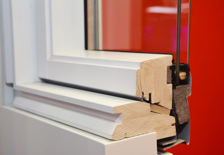 Esempio di Infissi e serramenti in legno: Vista ravvicinata di una sezione trasversale del telaio di una finestra che mostra l'interno in legno e l'esterno bianco, su uno sfondo rosso.